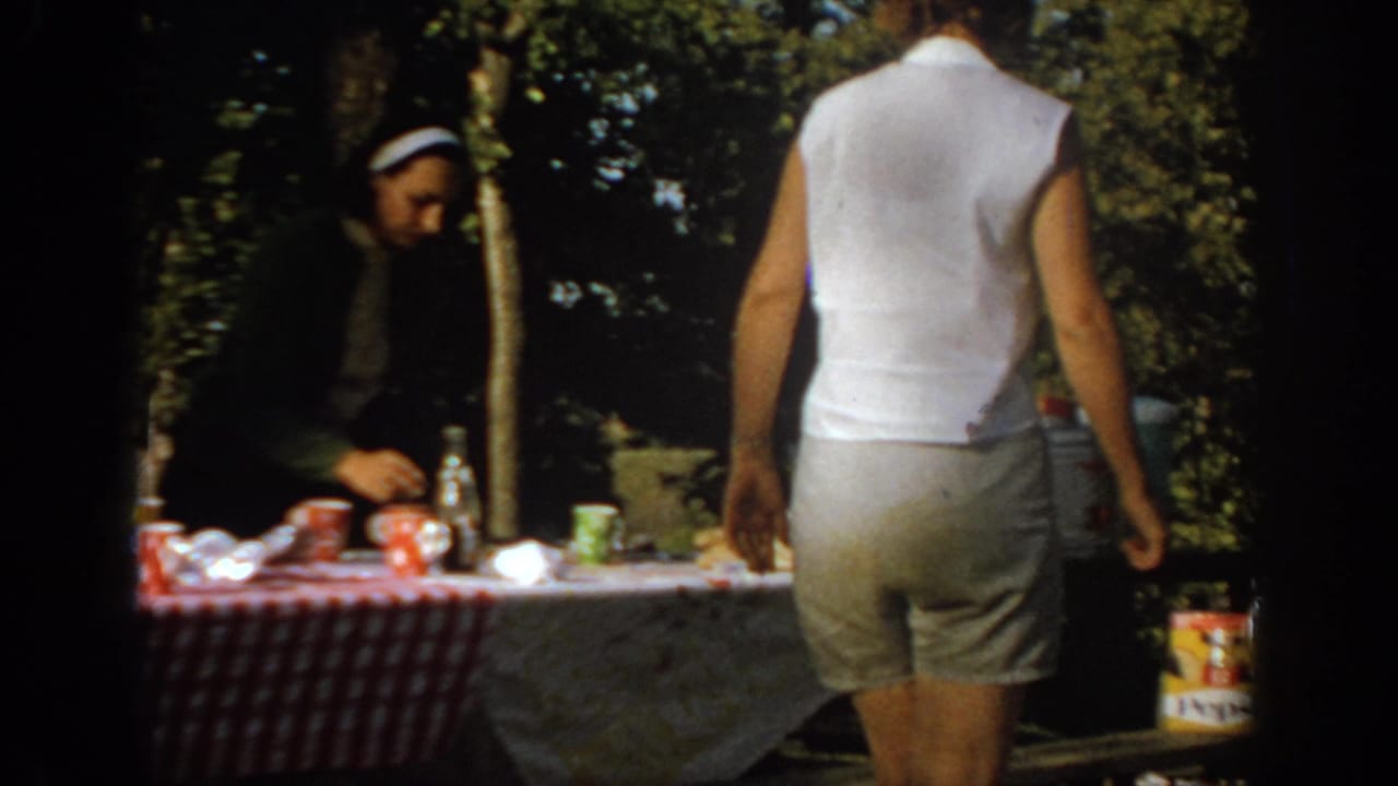 1967 Oklahoma home-movie frame showing adults near a backyard picnic table with a checkered cloth, food, drink containers, and shaded trees.