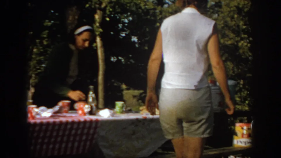 1967 Oklahoma home-movie frame showing adults near a backyard picnic table with a checkered cloth, food, drink containers, and shaded trees.
