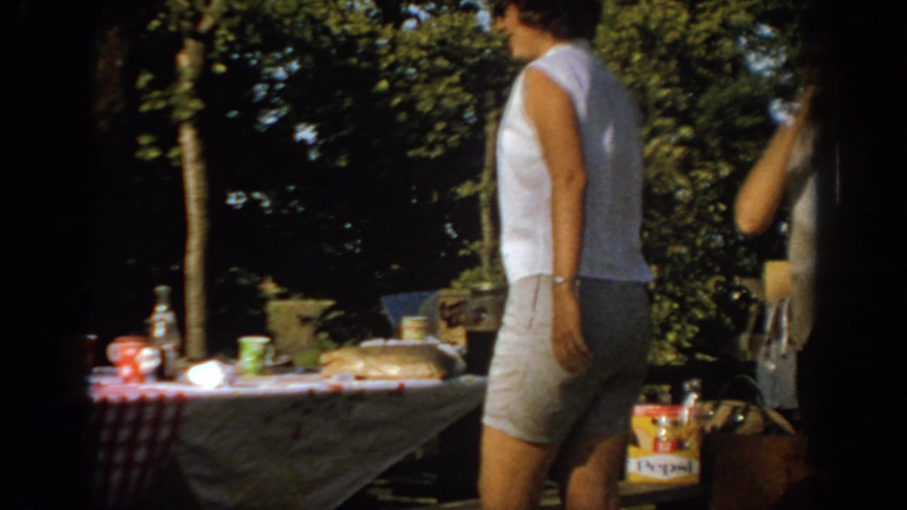 1967 Oklahoma backyard frame with a woman in a white sleeveless top near a picnic table holding food, bottles, and visible soft drink packaging.