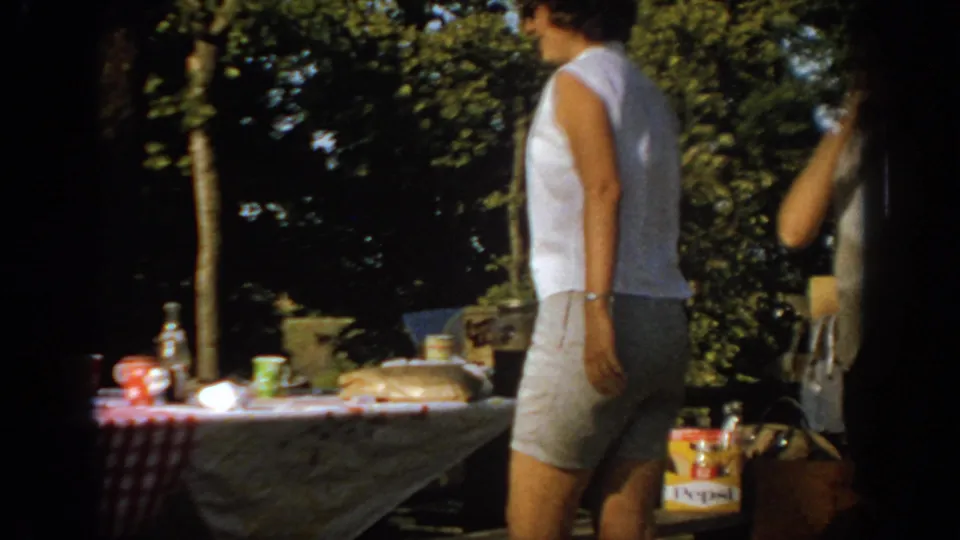 1967 Oklahoma backyard frame with a woman in a white sleeveless top near a picnic table holding food, bottles, and visible soft drink packaging.