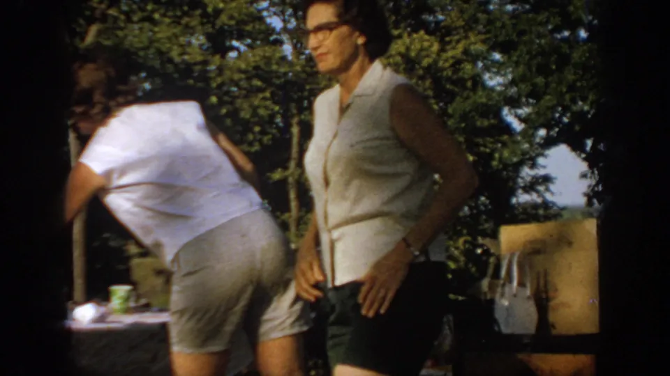 1967 Oklahoma home-movie frame showing two women beside a shaded backyard picnic table with cups, food, and trees in the background.