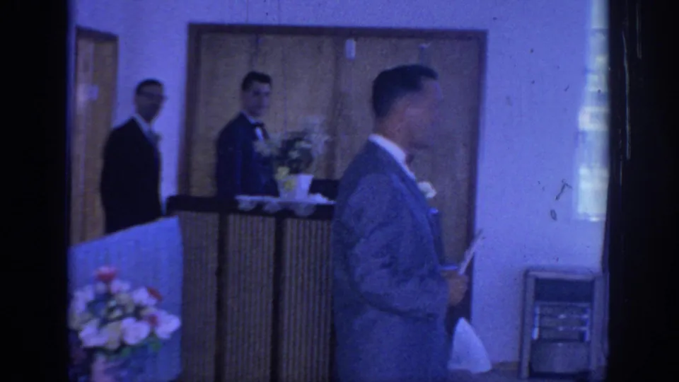 Indoor 1967 Oklahoma wedding-related frame with suited men, bow ties, a bouquet, wood doors, and blue-tinted 8mm interior color.