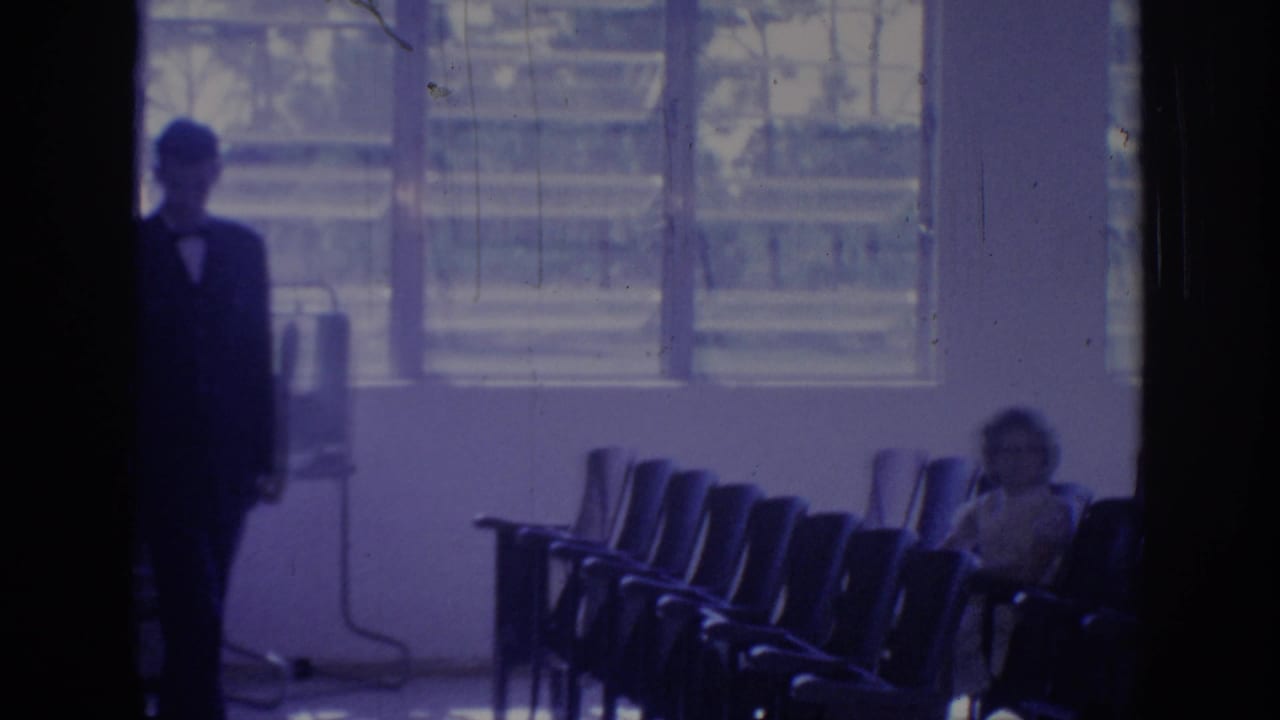 1967 Oklahoma 8mm frame showing a suited man near a dark doorway, rows of chairs, bright windows, and a seated woman.