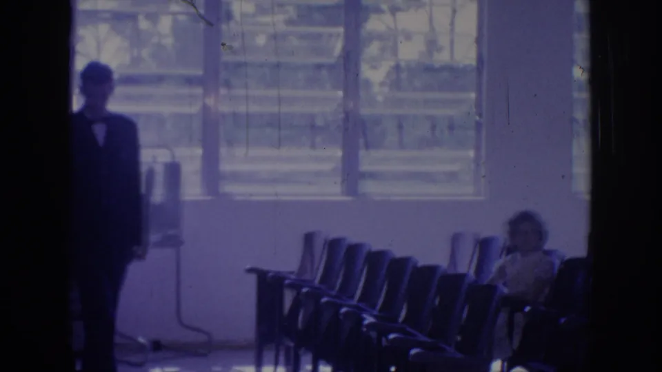 1967 Oklahoma 8mm frame showing a suited man near a dark doorway, rows of chairs, bright windows, and a seated woman.