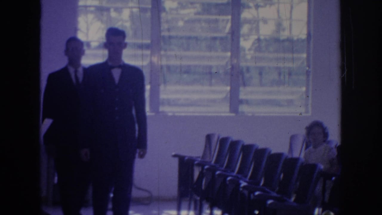 1967 Oklahoma 8mm frame with two men in dark suits, one wearing a bow tie, walking through a conference-style room.