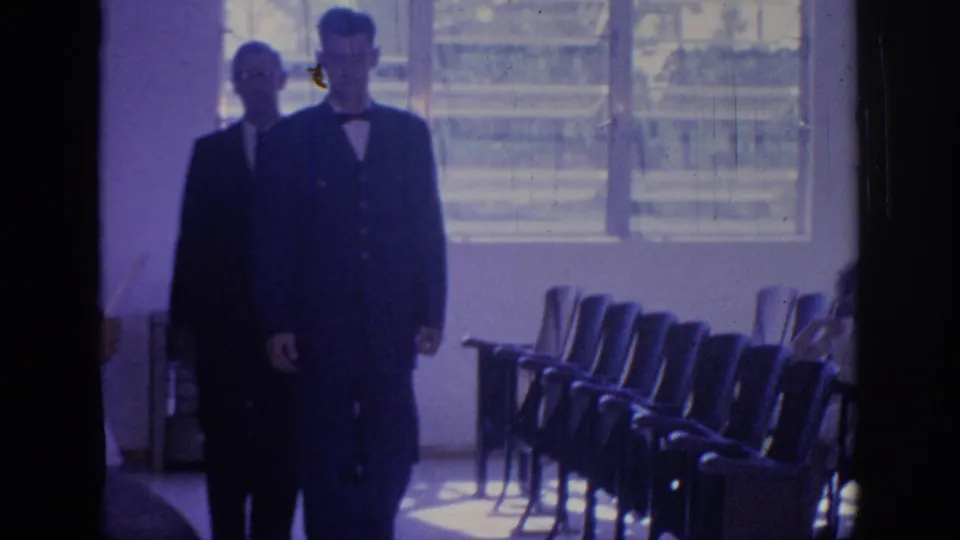 1967 Oklahoma 8mm frame showing two suited men close to the camera with chair rows, windows, and a partial seated figure nearby.