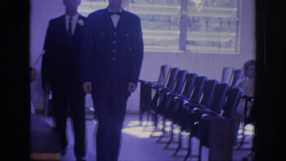 1967 Oklahoma 8mm frame with the suited men cropped close in front of rows of chairs and a seated woman near the wall.