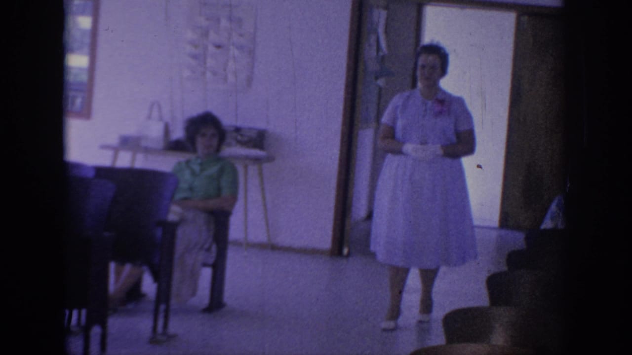 1967 Oklahoma home-movie frame showing a woman in a light dress walking inside a simple room with seated guests and chairs.