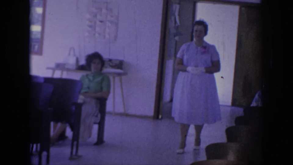 1967 Oklahoma home-movie frame showing a woman in a light dress walking inside a simple room with seated guests and chairs.