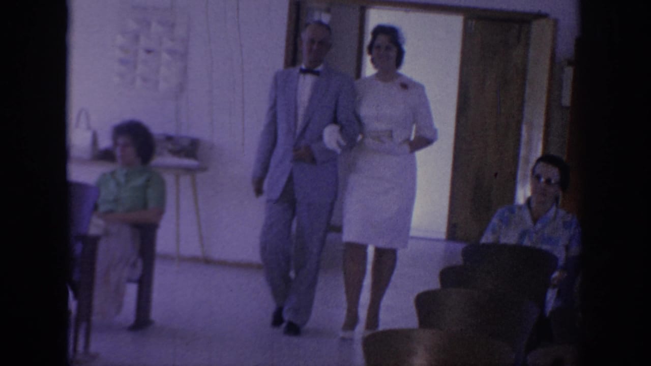 Man in a light suit walks arm in arm with a woman in a white dress past seated guests.