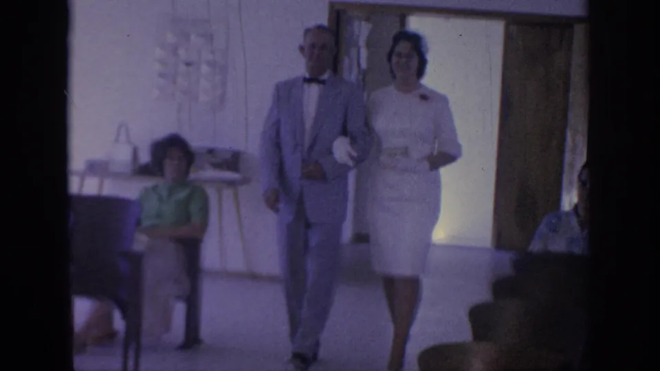 1967 Oklahoma home-movie frame showing the formally dressed couple nearer the camera in a plain indoor room with chairs and guests.