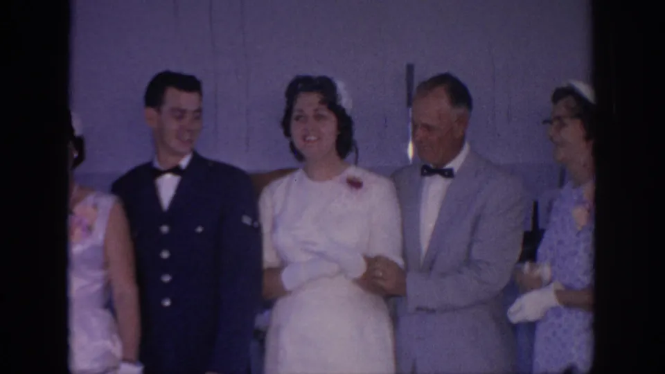 1967 Oklahoma wedding party stands indoors as the bride links arms with guests beside a groom in a dark formal uniform-style jacket.