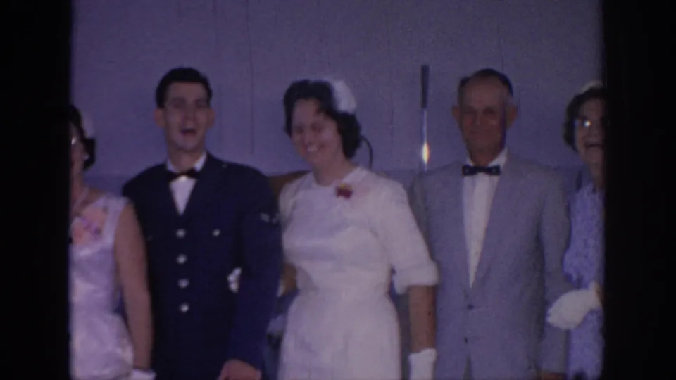 The bride and groom stand among formally dressed guests at a 1967 Oklahoma wedding, with faces visible and the group posed against a plain wall.