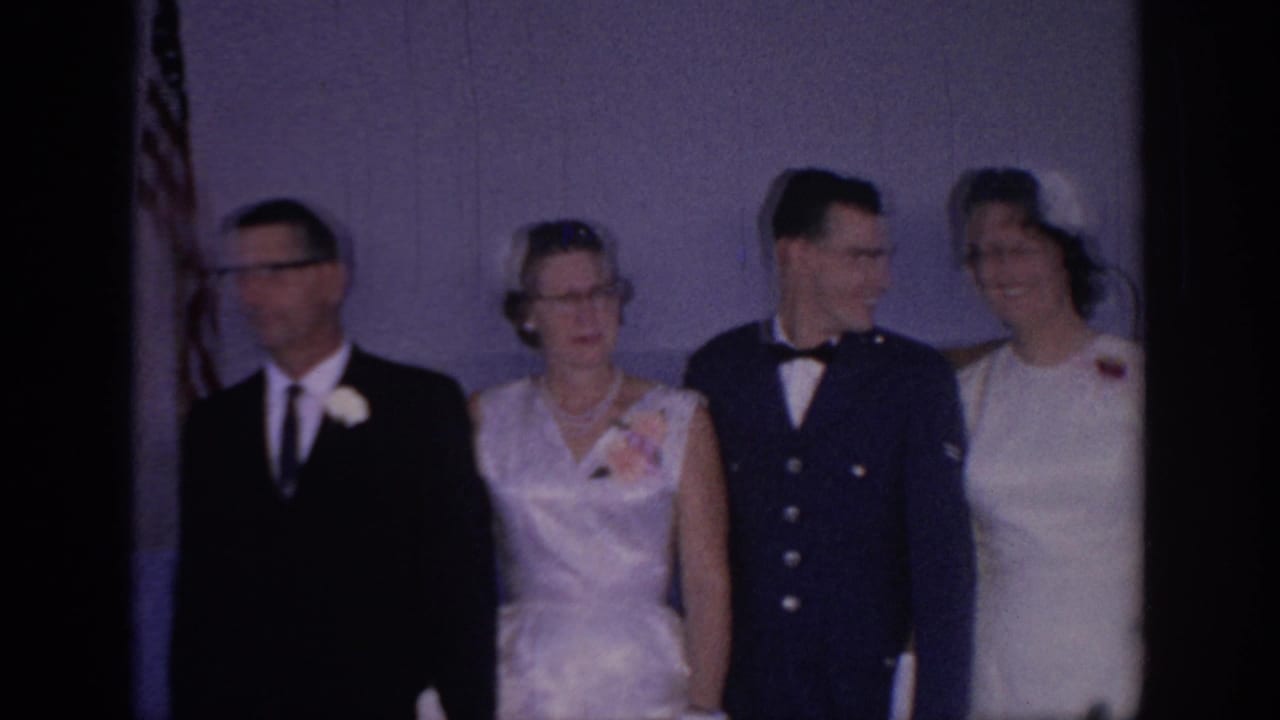1967 Oklahoma home-movie frame showing a bride, a groom in a dark military-style uniform, and older guests posing for wedding photos indoors.