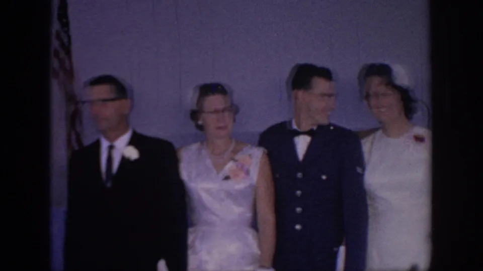 1967 Oklahoma home-movie frame showing a bride, a groom in a dark military-style uniform, and older guests posing for wedding photos indoors.