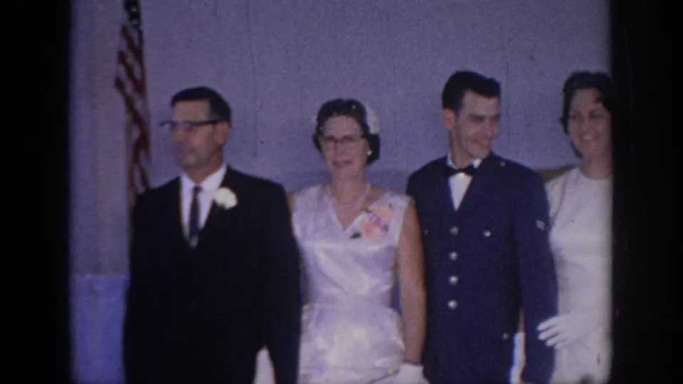1967 Oklahoma home-movie still of a bride, uniformed groom, and older relatives arranged for a wedding portrait near an American flag.