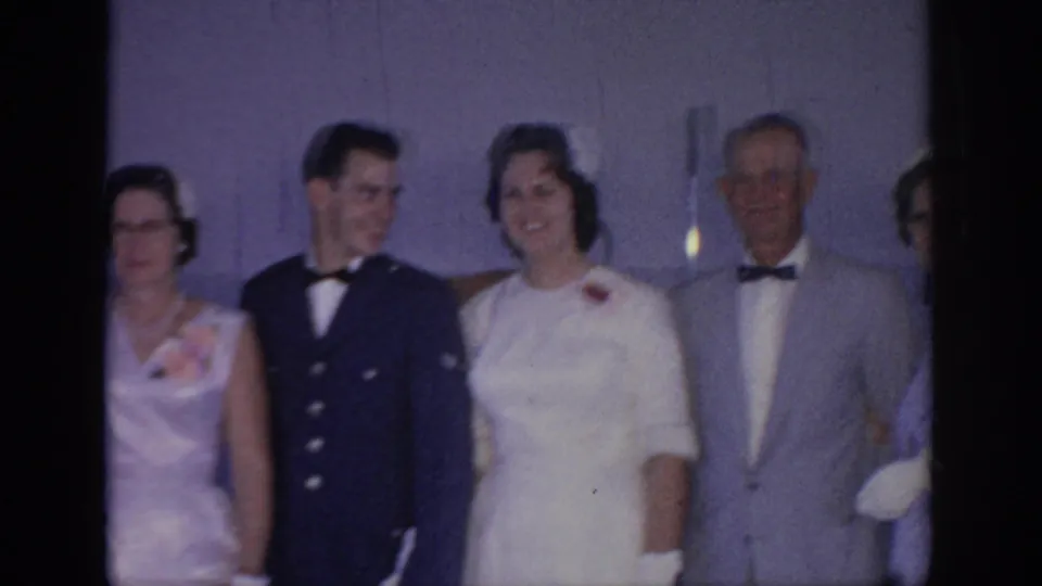 1967 Oklahoma wedding frame showing the bride and groom smiling with family members in formal clothing during an indoor portrait setup.
