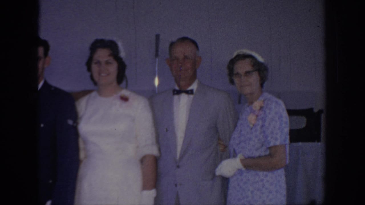 1967 Oklahoma home-movie frame showing a bride in white posing indoors with older relatives beside a purple-toned wall.