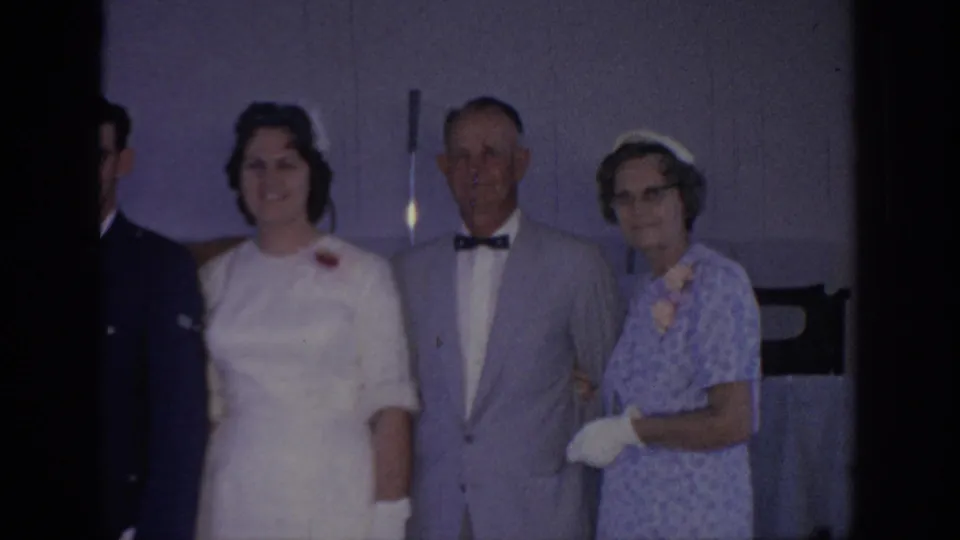 1967 Oklahoma home-movie frame showing a bride in white posing indoors with older relatives beside a purple-toned wall.