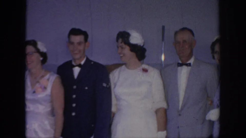 In this 1967 Oklahoma home-movie frame, the bride looks toward the uniformed man while older relatives remain lined up for the wedding portrait.