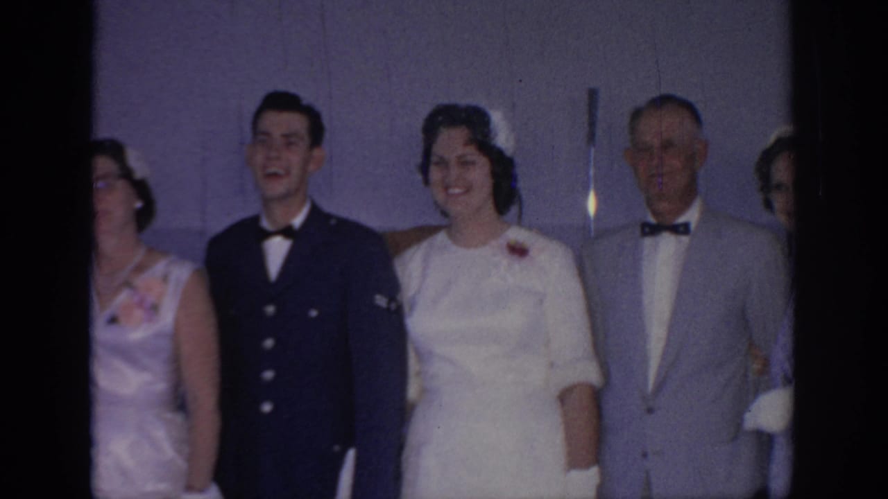 1967 Oklahoma home-movie frame of adults posed indoors at a formal gathering, with a uniformed man, a woman in white, and suited guests.