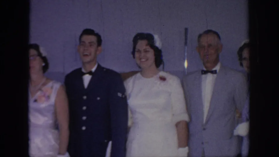 1967 Oklahoma home-movie frame of adults posed indoors at a formal gathering, with a uniformed man, a woman in white, and suited guests.