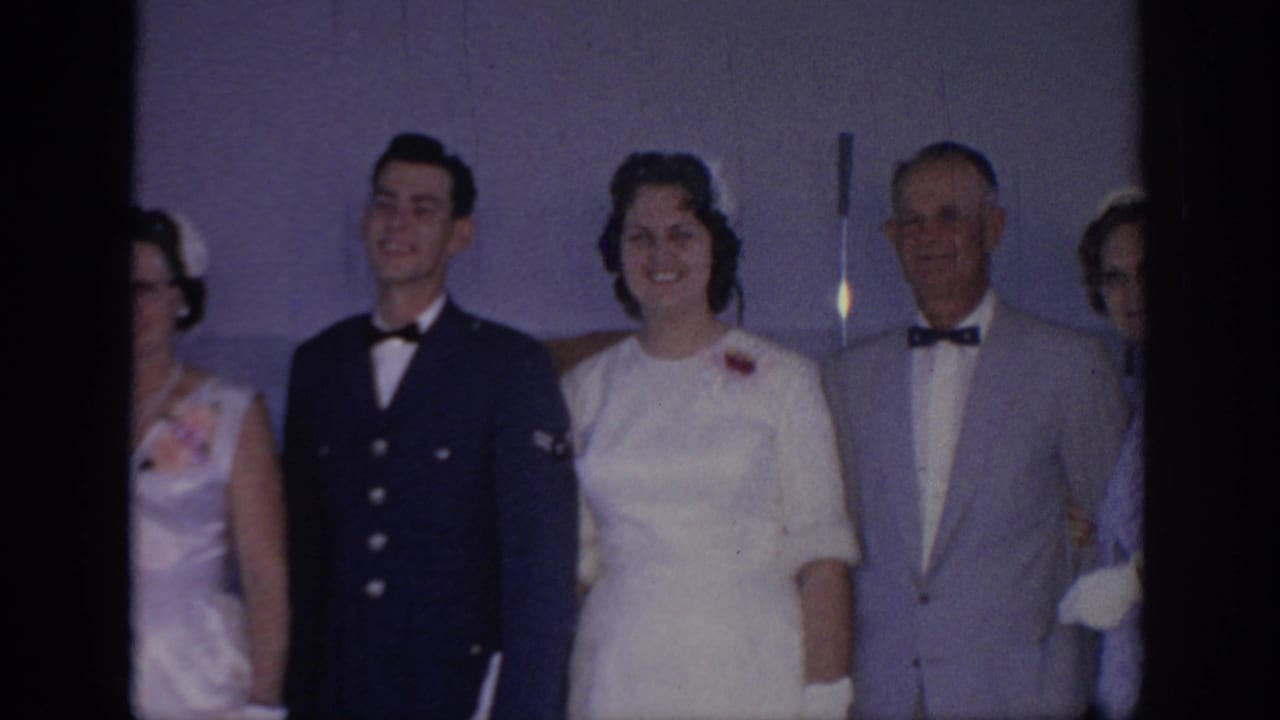 A uniformed man, woman in white, and older man in a light suit stand together indoors and smile.