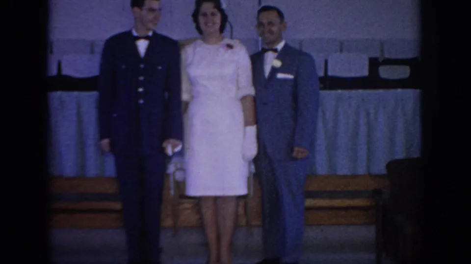 1967 Oklahoma home-movie frame of three formally dressed adults posed before banquet seating at an indoor social event.