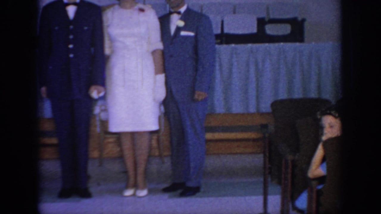1967 Oklahoma home-movie frame showing a bride, groom, and uniformed man standing before guests in a church or hall interior.