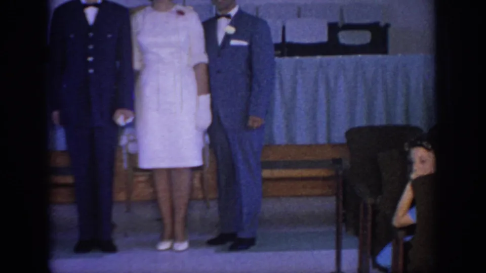 1967 Oklahoma home-movie frame showing a bride, groom, and uniformed man standing before guests in a church or hall interior.