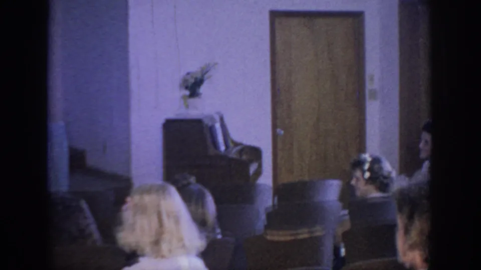 1967 Oklahoma home-movie frame from the guest area, showing attendees seated near a door and simple floral decoration.