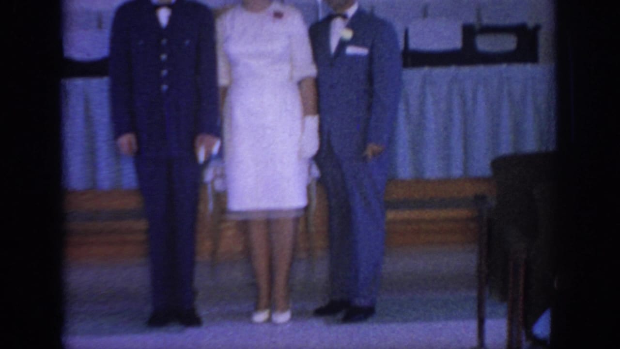 1967 Oklahoma home-movie frame showing three adults in formal wedding attire posed indoors, with a draped table and wooden steps behind them.