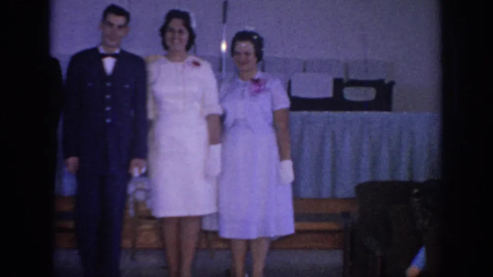 1967 Oklahoma wedding footage frame with a man in a dark military-style uniform beside two women in light formal dresses with corsages.