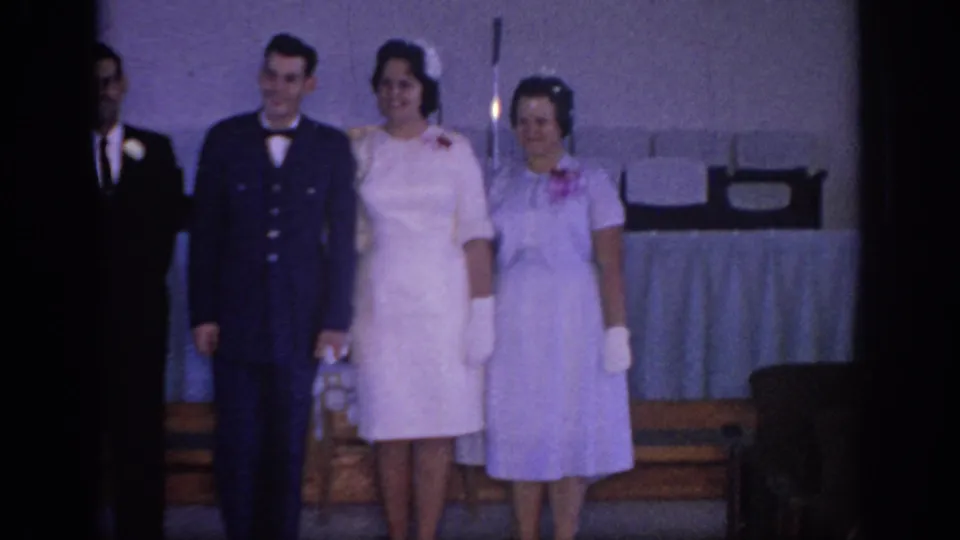 1967 Oklahoma home-movie frame showing four adults in formal wedding attire, including a dark suit, uniform, and light dresses, posed indoors.