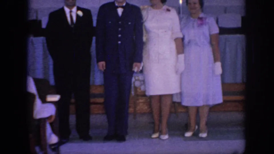 1967 Oklahoma wedding frame showing four adults posed on an indoor stage, with formal dresses, a dark uniform, wooden steps, and a draped table.