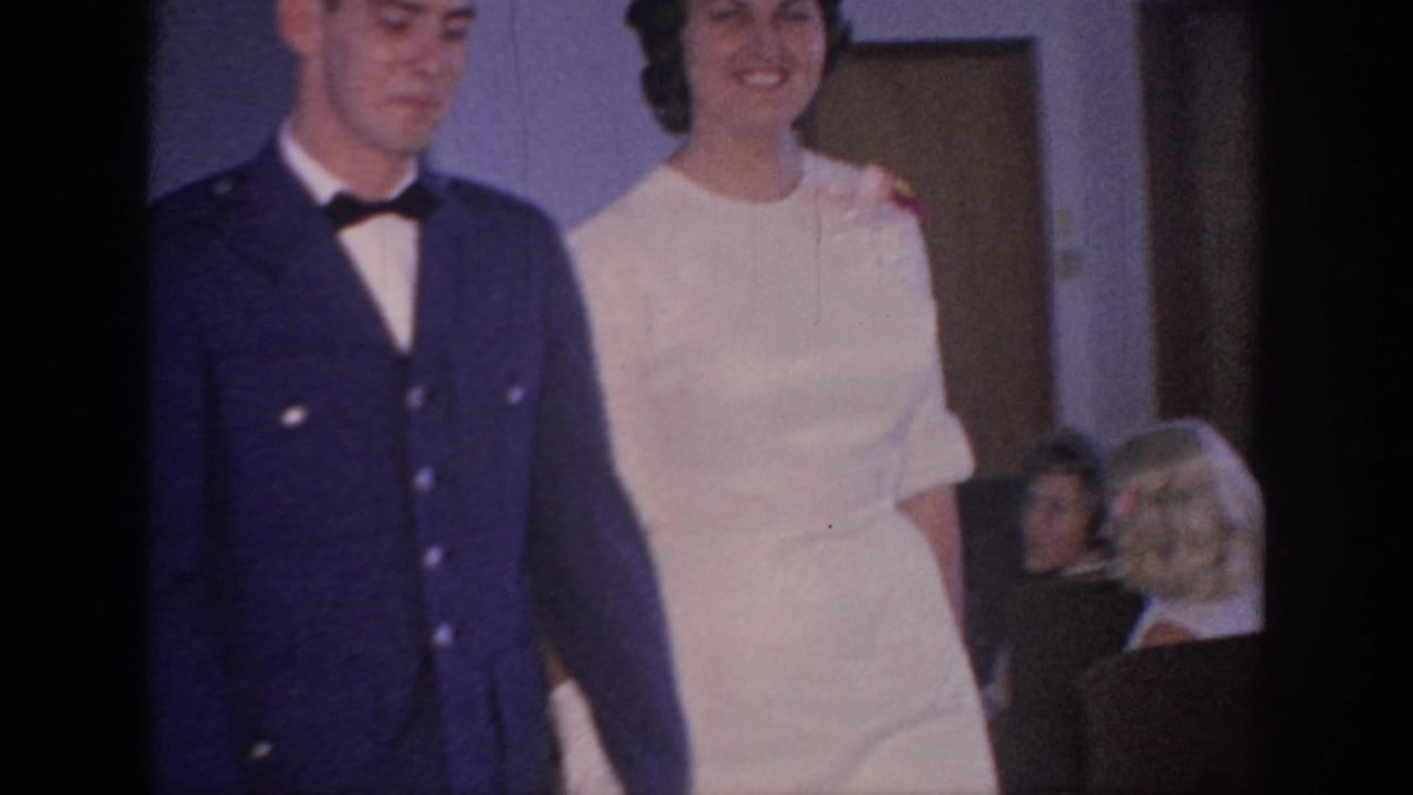 In 1967 Oklahoma home-movie footage, a man in a dark uniform walks beside a bride in white as seated guests watch indoors.