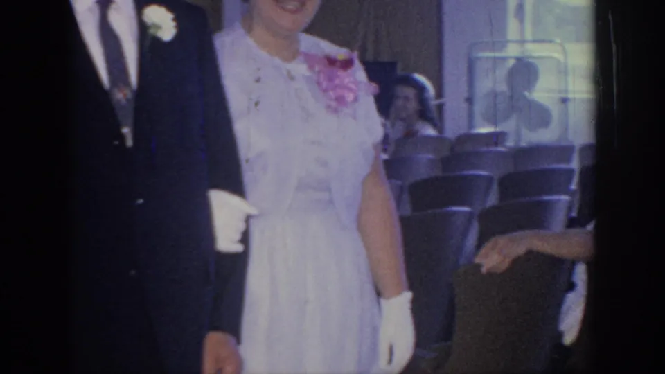 The camera catches a cropped view of a formal couple moving near rows of chairs, with another guest visible behind them.