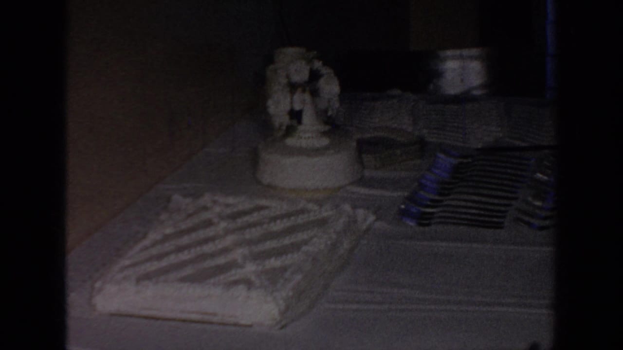 1967 Oklahoma home-movie frame showing a wedding cake table with a topper, plates, forks, and a tablecloth under dim indoor light.