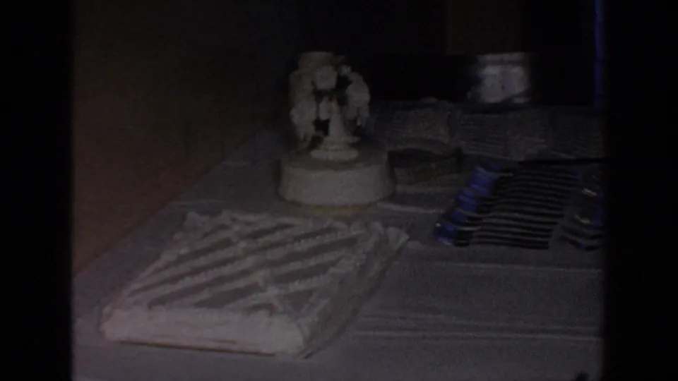 1967 Oklahoma home-movie frame showing a wedding cake table with a topper, plates, forks, and a tablecloth under dim indoor light.