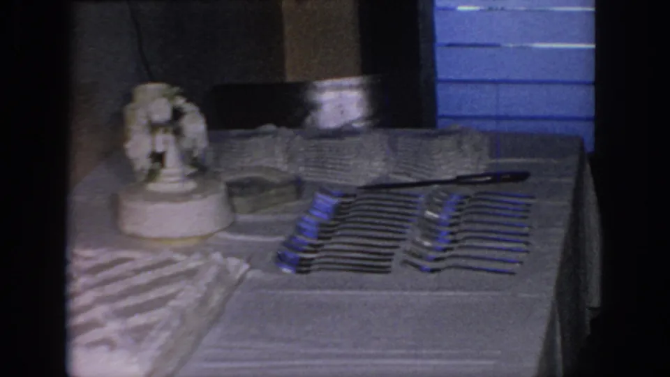 1967 Oklahoma home-movie view of the wedding reception table, showing the cake topper, stacked plates, arranged forks, and blue background panels.
