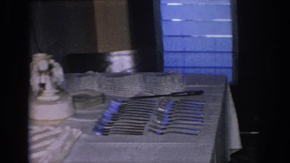 1967 Oklahoma frame of a wedding cake table with a figurine, stacked plates, rows of forks, a knife, and blue panels in the background.