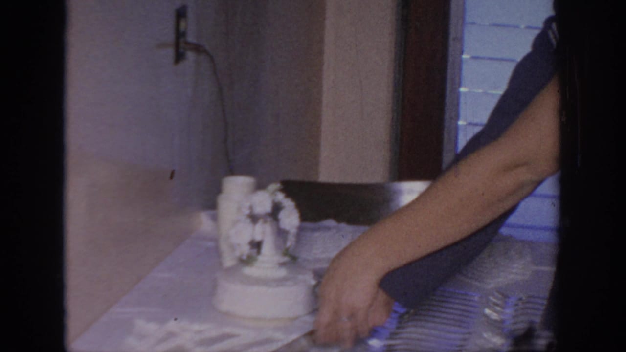 Arm reaching toward a white frosted cake on a table with a small ornament.