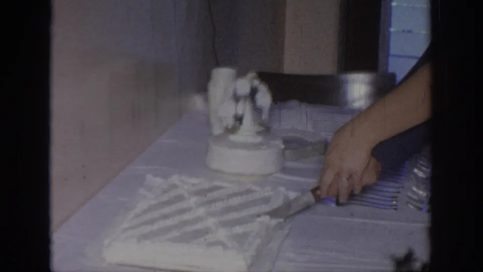 A hand cuts into the white frosted cake with a knife during the supported 1967 Oklahoma home-movie celebration.