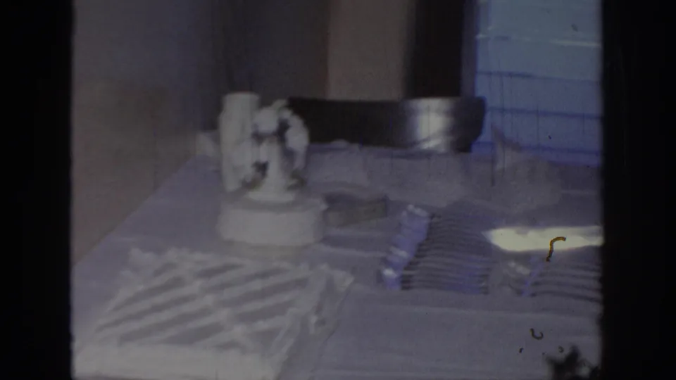 1967 Oklahoma 8mm frame showing a white frosted wedding cake, stacked forks, and table setting in an indoor reception space.