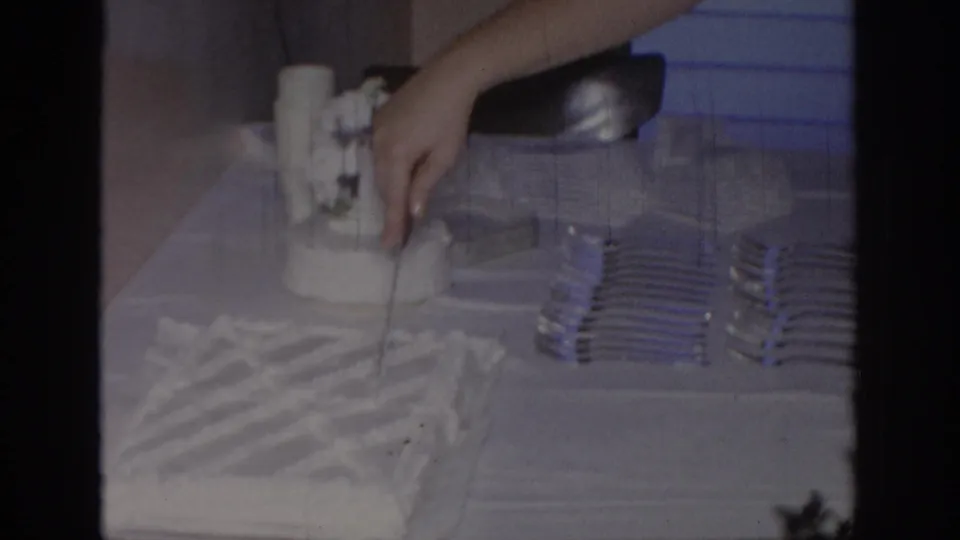A close 1967 Oklahoma home-movie view shows a hand reaching to cut the white frosted cake beside stacked forks and blinds.