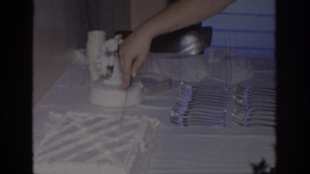 1967 Oklahoma home-movie frame showing an adult hand near a white frosted cake beside stacked forks, napkins, and a tablecloth.
