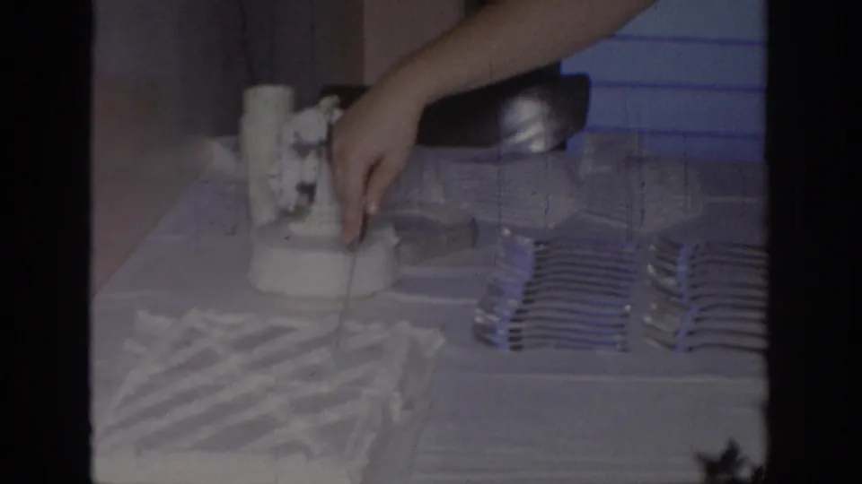 1967 Oklahoma home-movie frame showing an adult hand near a white frosted cake beside stacked forks, napkins, and a tablecloth.