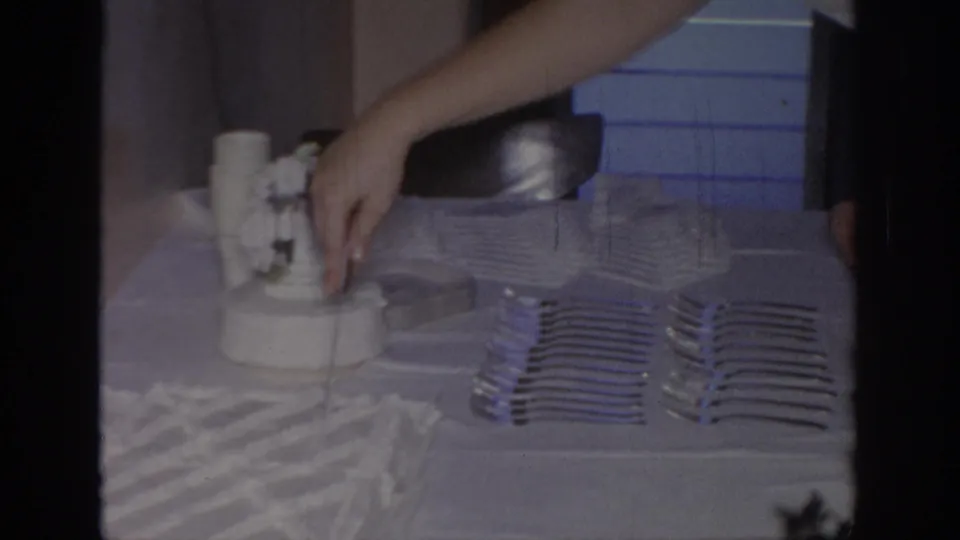 1960s home-movie frame of a hand cutting into a white frosted cake on an indoor table set with forks and napkins.