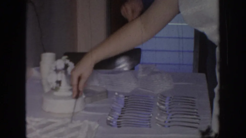Adult arm and partial torso lean over a celebration table in Oklahoma footage, cutting or serving a white frosted cake.