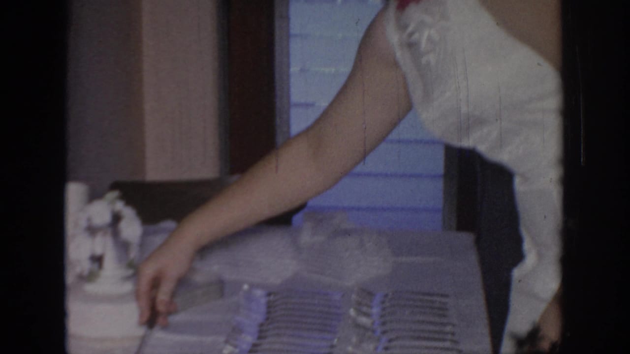 In a 1967 Oklahoma home-movie frame, a woman in a white sleeveless dress appears to arrange tableware on a serving surface.